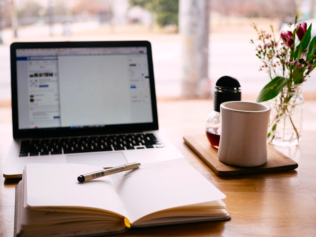 Open notebook in front of a notebook computer on a desk with flowers in a vase